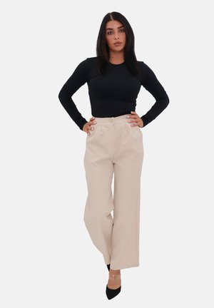 Schwarzes langärmliges Oberteil, kombiniert mit hochtaillierten, weit geschnittenen beigen Hosen. Die Hose hat einen Frontknopf und eine glatte Textur. Schwarze Absätze runden das Outfit ab.