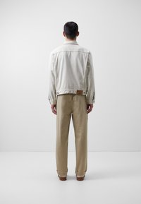 Mand står med ryggen til, iført en lys denimjakke og beige bukser med opfoldede ben i et enkelt hvidt rum.