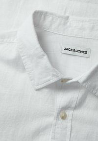 Camisa de cuello blanco con una tela texturizada. Presenta una etiqueta de marca, un solo botón y una costura limpia a lo largo del cuello y la tapeta.