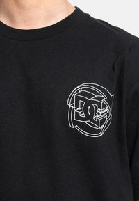 Black cotton t-shirt with a round neckline, featuring a white graphic design of the DC logo on the left chest area.