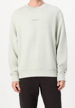 Mann trägt ein hellgraues, strukturiertes Langarm-Sweatshirt mit dem "tommy jeans" Logo und schwarze Hosen vor weißem Hintergrund.