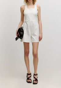 Witte katoenen romper met brede bandjes en een getailleerd lijfje. Losse shorts en zichtbare naden; gecombineerd met zwarte sandaaltjes met banden en een clutch.
