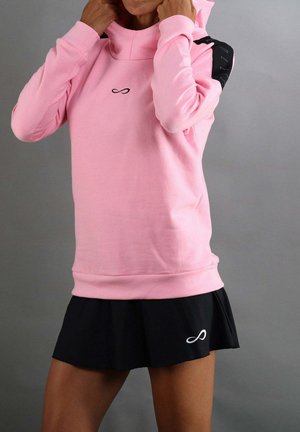 Persoon die een roze hoodie en een zwarte rok met oneindigheidssymbolen draagt, en de capuchon boord aanpast tegen een effen grijze achtergrond.