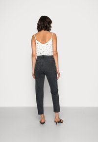 Donna con capelli corti scuri, che indossa una canotta bianca a fiori, jeans neri aderenti a vita alta e sandali con tacco neri, in piedi contro un muro bianco.