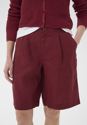 Kobieta nosząca burgundowe lniane szorty z wysokim stanem, luźnym krojem, zakładkami z przodu i bocznymi kieszeniami, zestawione z bordowym dzianinowym kardiganem i białą koszulą.