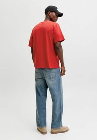 Punane puuvillane T-särk lühikeste varrukatega, koos helesiniste laiade teksadega. Mudel kannab musta mütsi ja beeže kingi.