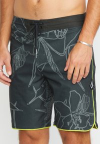 Pantaloni da bagno neri con motivi floreali bianchi, bordi verdi a contrasto, vita con cordino regolabile e logo sul lato.