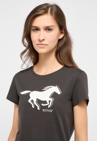 Tmavě šedé tričko s krátkým rukávem s bílým grafickým potiskem skákajícího koně a logem "MUSTANG" níže. Měkká látka s klasickým kulatým výstřihem.