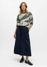 Kvinde iført grå og hvid mønstret sweater, mørkeblå lang denimnederdel og sorte loafers, stående med den ene hånd i lommen.