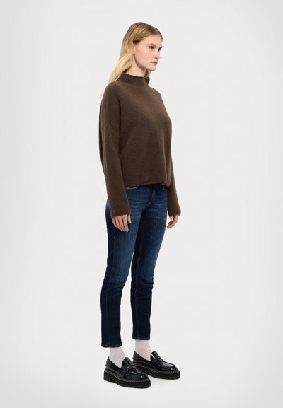 Brązowy sweter z dzianiny z wysokim kołnierzem, w połączeniu z ciemnoniebieskimi dżinsami i czarnymi mokasynami. Model stoi na tle jasnego, gładkiego tła.