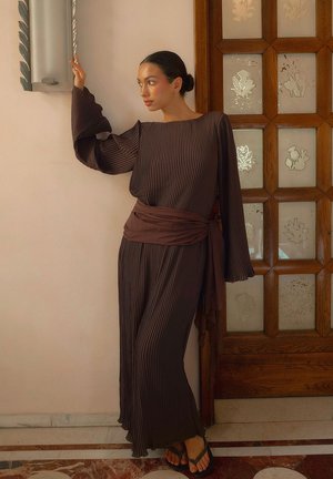 Robe plissée marron chocolat avec des manches évasées et une ceinture assortie, accentuée par une texture lisse, portée avec des tongs noires.