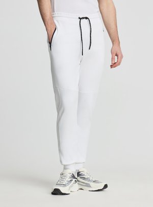 Uomo che indossa pantaloni da jogging bianchi con coulisse nera e tasche con cerniera, abbinati a scarpe sportive bianche e grigie, in piedi su sfondo semplice.