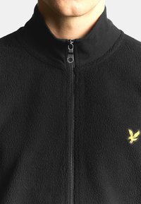 Schwarze Fleecejacke mit Reißverschluss, hohem Kragen, strukturierter Oberfläche und kleinem gelbem, gesticktem Logo auf der linken Brust.