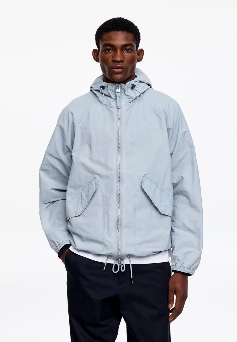 Mand iført lys grå hættejakke med frontlynlås og store klaplommer, stående med hænderne i lommerne mod en hvid baggrund.