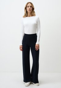 Un haut à manches longues blanc et un pantalon large à taille haute de couleur bleu marine foncé avec des coutures frontales, associés à des baskets blanches sur un fond uni.