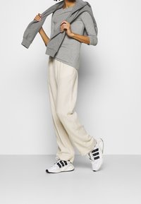 Grijze longsleeve top, crème wijde broek en witte sneakers met zwarte strepen. Een grijze sweatshirt over de schouders gedrapeerd.