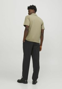 Polo shirt khaki a maniche corte con una texture morbida, vestibilità rilassata e ventole laterali, abbinato a pantaloni neri e scarpe nere.