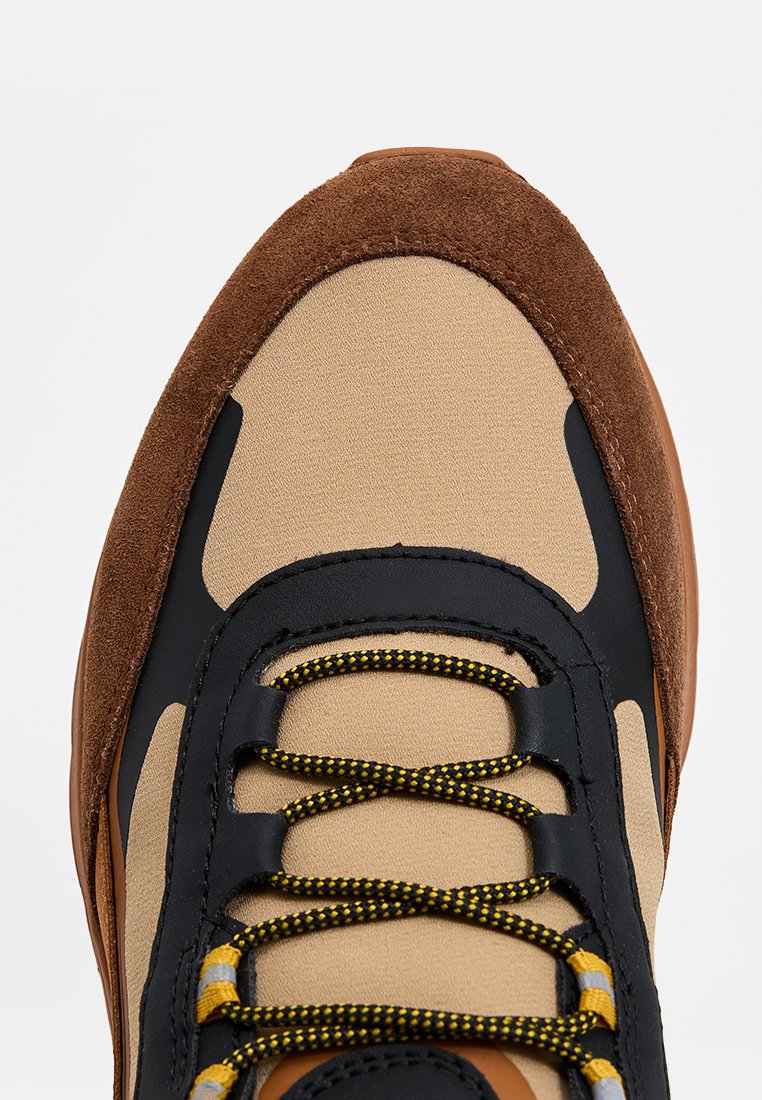 Baskets beiges et brunes avec une tige texturée, des accents noirs et des lacets jaunes. Talon en daim et matériau en mesh respirant.