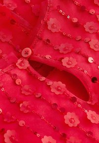 Tissu rouge transparent orné d'applications florales et de paillettes, avec une fermeture à boutons. Surface texturée avec des embellissements alternés.