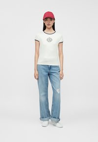 Νεαρή γυναίκα που φοράει κόκκινο καπέλο, λευκό εφαρμοστό μπλουζάκι με λογότυπο, ανοιχτό μπλε τζιν παντελόνι με τρύπα και λευκά αθλητικά παπούτσια, στέκεται μπροστά σε απλό φόντο.