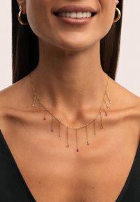 Collier en chaîne dorée avec plusieurs petits charms pendants en pierres précieuses colorées, porté avec des boucles d'oreilles créoles dorées et un col de haut noir.