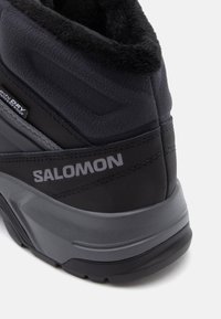 Czarne wodoodporne buty z teksturowanej tkaniny, z wyściółką przy kołnierzu i szczegółowym szarym logo. Wzmocniona podeszwa zapewniająca przyczepność.