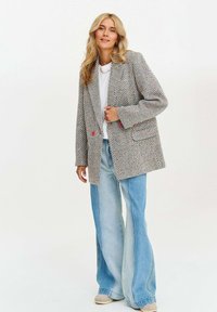 Žena s dlouhými blond vlasy, oblečená v texturovaném oversized saku, bílém topu, širokých dvoubarevných modrých džínech a béžových botách, stojící na bílém pozadí.