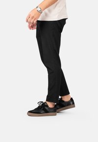 Pantalones de algodón negros con un corte ajustado, combinados con zapatillas negras que tienen una superficie texturizada y suela marrón. El reloj es de metal plateado con una caja cuadrada.