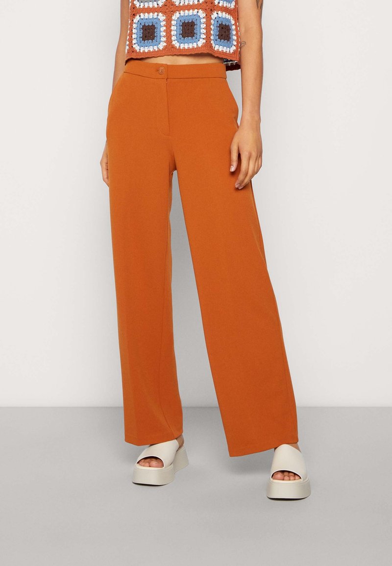 Femme portant un pantalon large orange, un top court avec un motif au crochet bleu, marron et blanc, et des sandales blanches à plateforme.