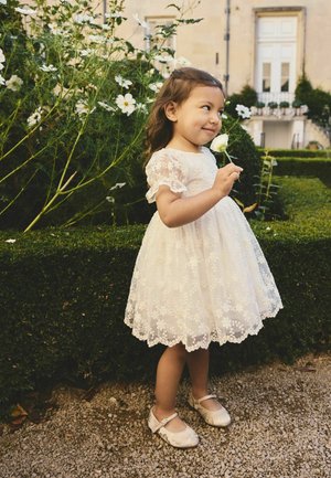 Robe en dentelle blanche avec motifs floraux, manches courtes à volants et ourlet festonné. Ballerines marron avec une bride. Enfant tenant une rose.