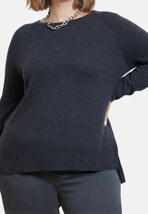 Frau trägt einen dunkelgrauen Langarmpullover und passende Hose mit einer silbernen Halskette, Hand ruht auf der Hüfte.