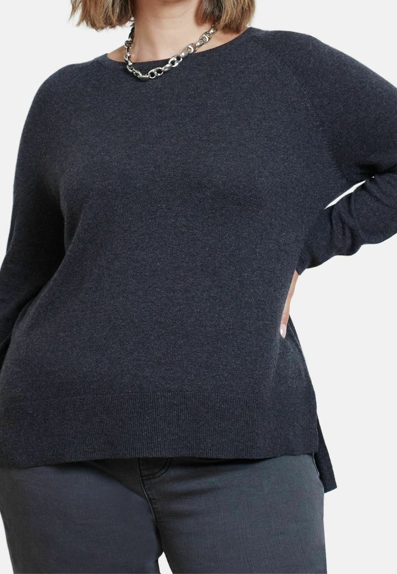 Femme portant un pull à manches longues gris foncé et un pantalon assorti, avec une chaîne en argent au cou, main posée sur la hanche.