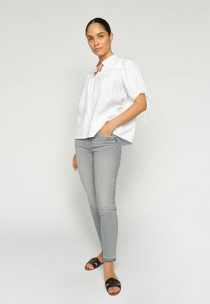 Naine seisab, kandes lahtist valget pluusi puffy varrukatega, helehallid kitsad teksapüksid ja mustad avatud varvastega sandaalid, lihtsalt taustal.