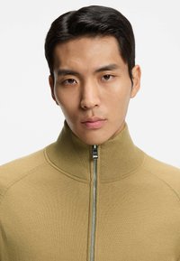 Beżowy sweter na zamek błyskawiczny z wysokim kołnierzem, prążkowaną fakturą i srebrnym zamkiem z przodu. Gładka tkanina z minimalną ilością detali.