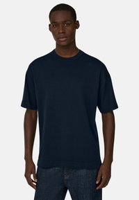 Jeune homme portant un t-shirt oversize uni bleu marine foncé et un jean bleu foncé, debout devant un fond gris clair.