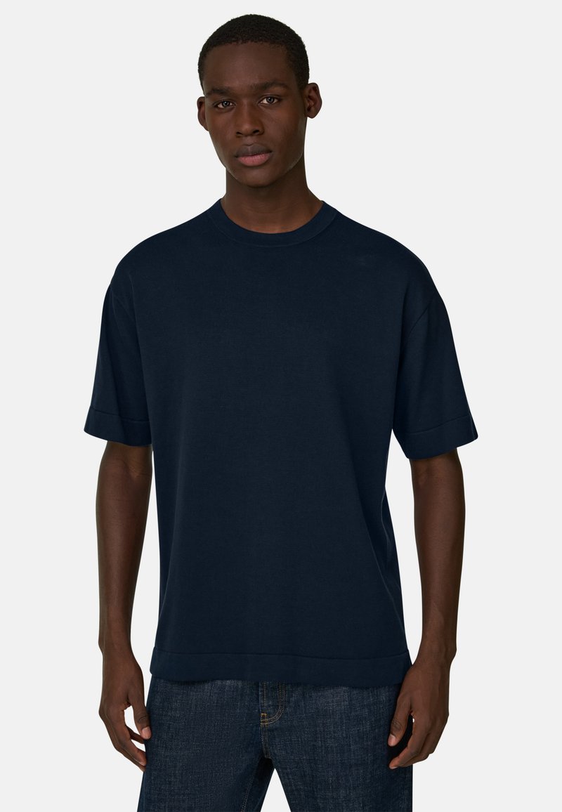 Jeune homme portant un t-shirt oversize uni bleu marine foncé et un jean bleu foncé, debout devant un fond gris clair.