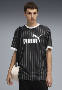 Tricou cu dungi negre și accente albe, având un mare logo "PUMA" pe față. Modelul ține o geantă de culoare crem, cu curea de umăr.