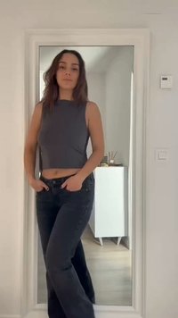 Mujer con cabello largo y oscuro, de pie con las manos en los bolsillos, vistiendo una camiseta sin mangas oscura y jeans frente a un espejo de cuerpo entero.