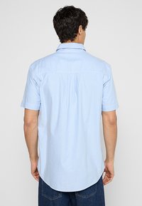Camicia azzurra a maniche corte con colletto a punta, dettaglio della piega posteriore e orlo curvo. Realizzata in un tessuto morbido e strutturato.
