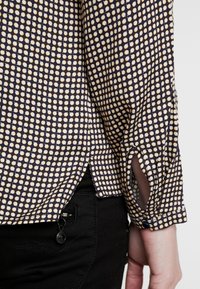 Ärmlinning och sidokant på en marinblå skjorta med vit och gul prickmönster, kombinerad med svarta byxor och synlig hand vid sidan.