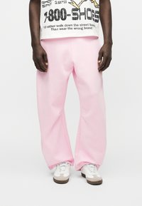 Άνδρας που φοράει φαρδιά ροζ παντελόνια, λευκό μπλουζάκι με τύπωμα και λευκά αθλητικά παπούτσια, στέκεται μπροστά σε απλό φωτεινό φόντο.