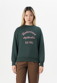 Zelená mikina so zvinutými manžetami a spodným okrajom, s ružovým vyšitým textom: "Timberland Authentic Est 1973". Nosená s hnedými nohavicami.