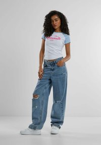 Camiseta blanca con mangas azul claro y gráfico rosa, combinada con vaqueros de denim azul holgados con rotos, y zapatillas blancas.