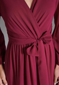 Burgunderfarbene Wickelkleid mit tiefem V-Ausschnitt, langen Ärmeln, strukturierter Stoff und einem gebundenen Taillendetails. Mit gerafften Falten von der Taille.
