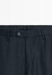 Linea della vita di pantaloni eleganti blu navy con chiusura a bottone, passanti per cintura e dettaglio in tessuto strutturato su sfondo bianco.