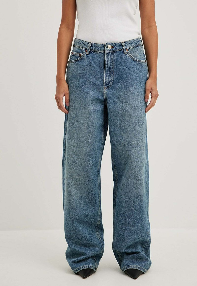 Šviesių džinsų plačios kojos antblauzdžiai su aukštu liemeniu, turintys klasikinį penkių kišenių dizainą, metalinį užsegimą ir švelnų blukimą.