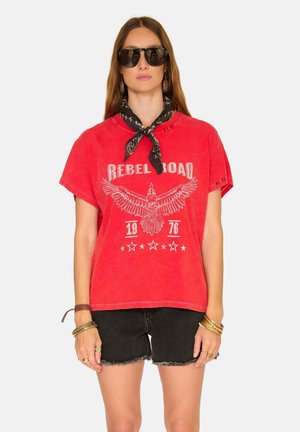 Femme portant un t-shirt rouge surdimensionné avec l'inscription "Rebel Road 1976", une écharpe noire nouée au cou, un short en denim noir, de grandes lunettes de soleil et des bracelets dorés.