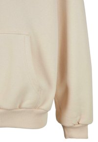 Felpa beige chiaro con tasca frontale, polsini e orlo a coste, realizzata in tessuto morbido. La texture appare liscia e priva di decorazioni.