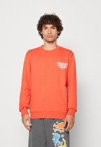 Felpa arancione con collo a giro e maniche lunghe, caratterizzata da un logo bianco "TOMMY JEANS" sul lato sinistro del petto. Tessuto morbido e texturizzato.
