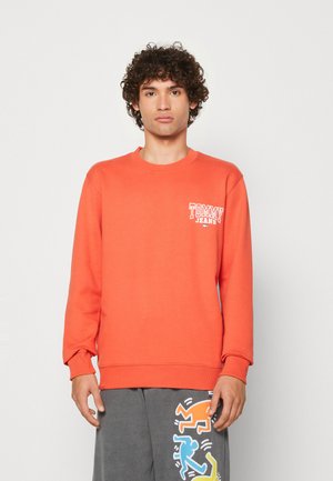 Felpa arancione con collo a giro e maniche lunghe, caratterizzata da un logo bianco "TOMMY JEANS" sul lato sinistro del petto. Tessuto morbido e texturizzato.
